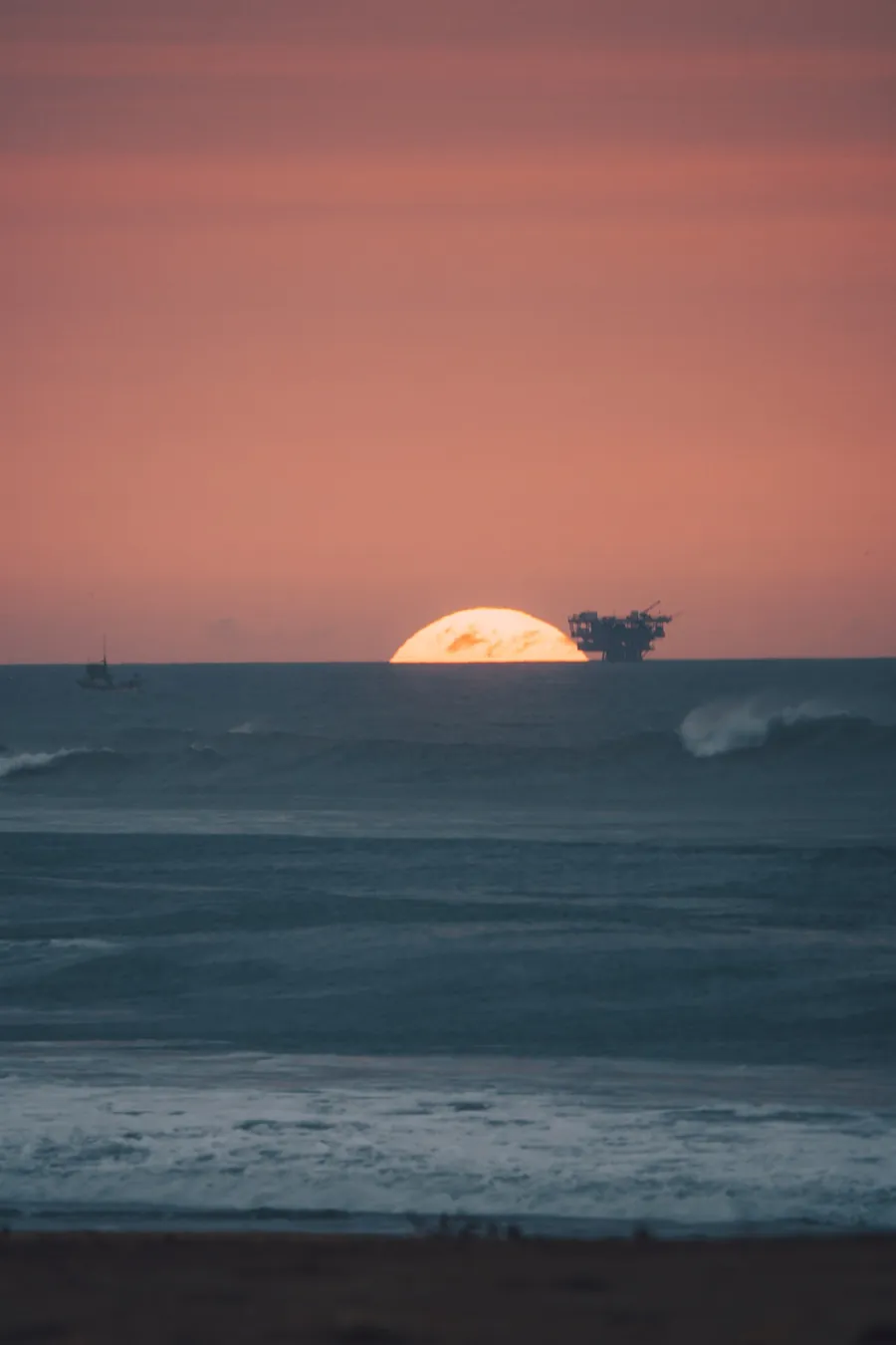 Sol se pondo no horizonte do mar, com um barco de pesca e uma plataforma ao longe, sob o céu rosado.