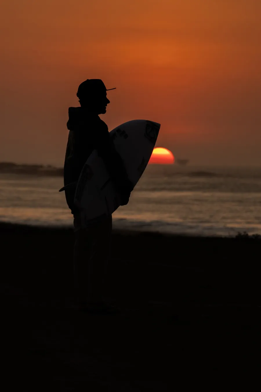 Silhueta de surfista com prancha debaixo do braço, com o sol se pondo no mar ao fundo.